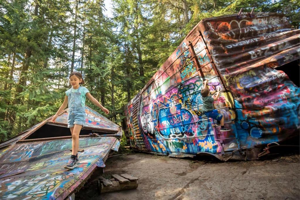 Two children exploring the colorful, graffiti-covered boxcars of the Whistler Train Wreck, nestled in a forest of tall evergreens on a sunny summer day.