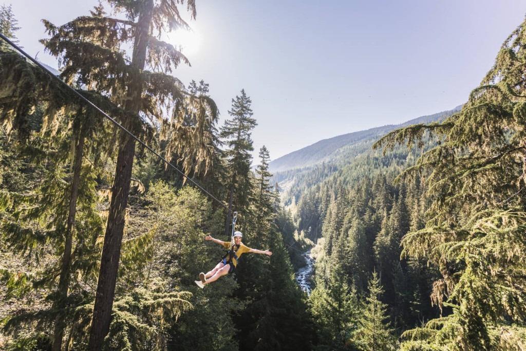 Thrill-seeker zip-lining through a lush forest with mountain views in Whistler, BC, arms outstretched under clear blue skies, capturing the excitement of outdoor adventure in the Coast Mountains.